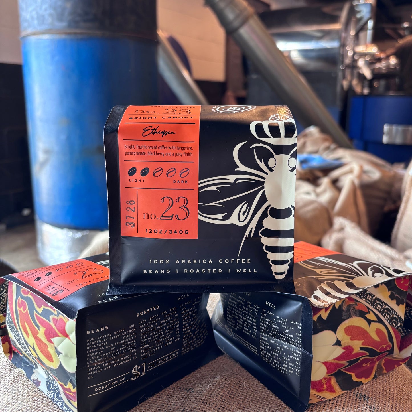 Three bags of coffee beans arranged on a burlap surface, with the central bag standing upright and showing a black package with a white bee illustration and orange label. The background shows a rustic coffee-roasting environment with a blue metal container and machinery.