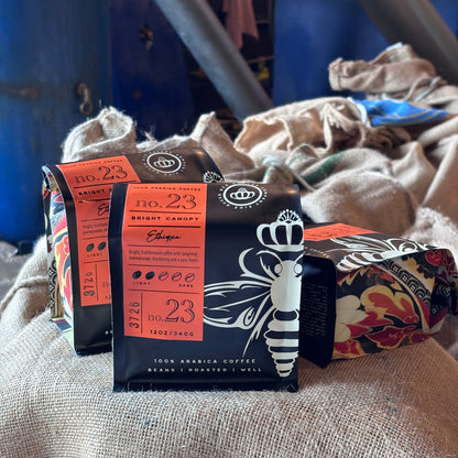 Three packaged bags of coffee stacked on top of each other, featuring dark navy and orange packaging with a white bee illustration, placed on a beige woven fabric surface. 